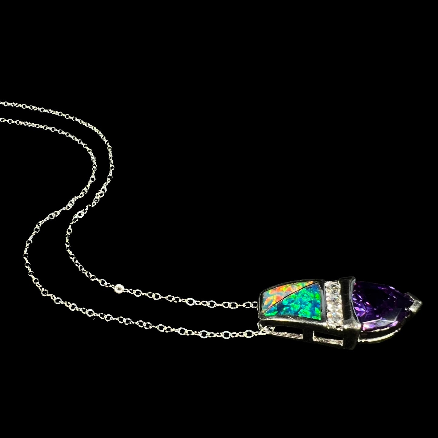 Angled side view of a sterling silver lab-created opal inlay and purple trillion cut CZ drop necklace, laying on a black background with its chain flowing behind the pendant.