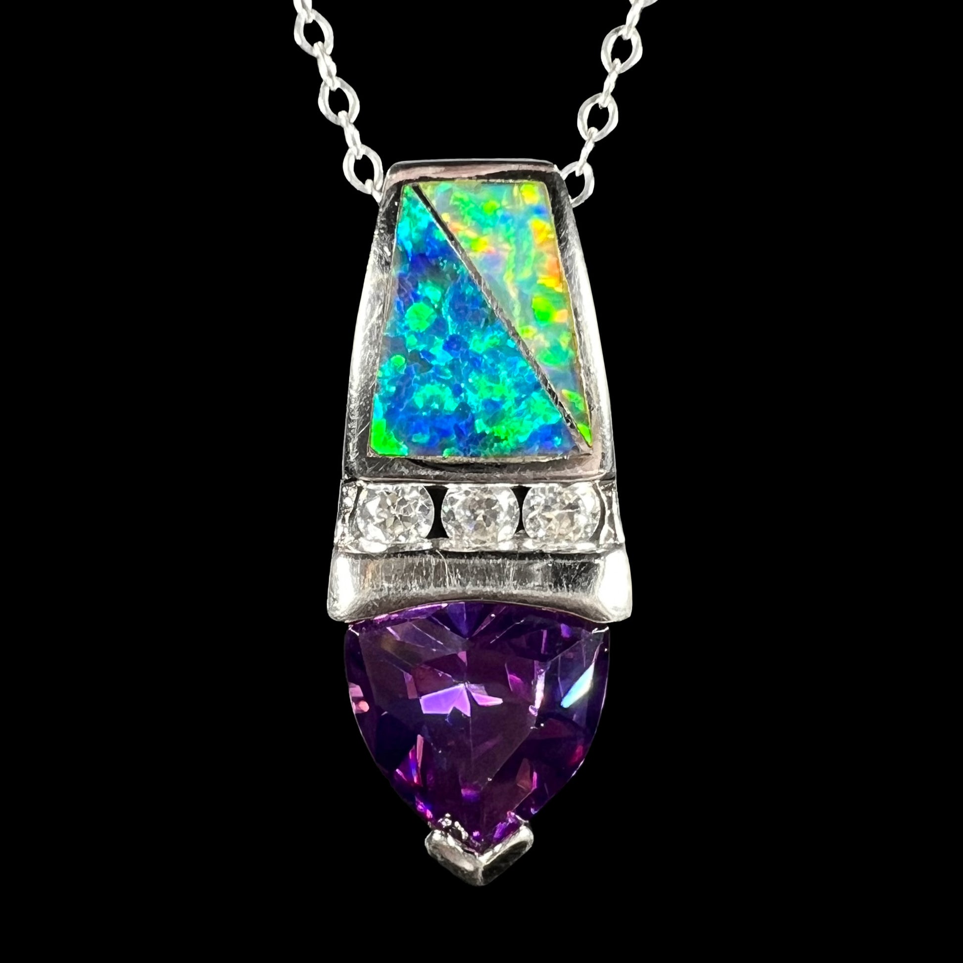 A sterling silver drop necklace featuring two lab-created opal inlay pieces, a row of colorless round cubic zirconia, and a trillion cut purple CZ, shown close-up on black background.