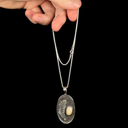 A sterling silver men's fire opal necklace by Austin Henderson, dangled by a hand from its box chain against a black background.