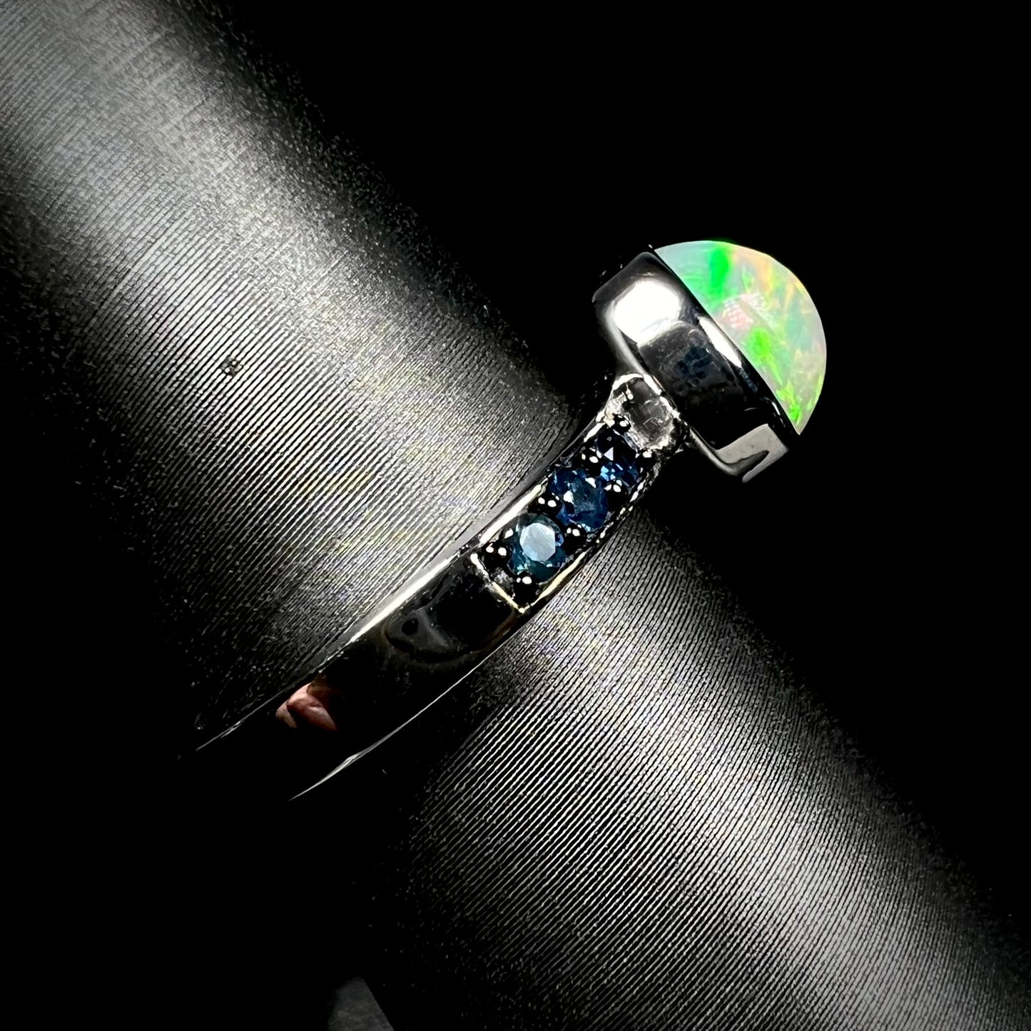 Side view of Ethiopian opal ring.  Channel set topaz accents and silver shank in focus against black background.