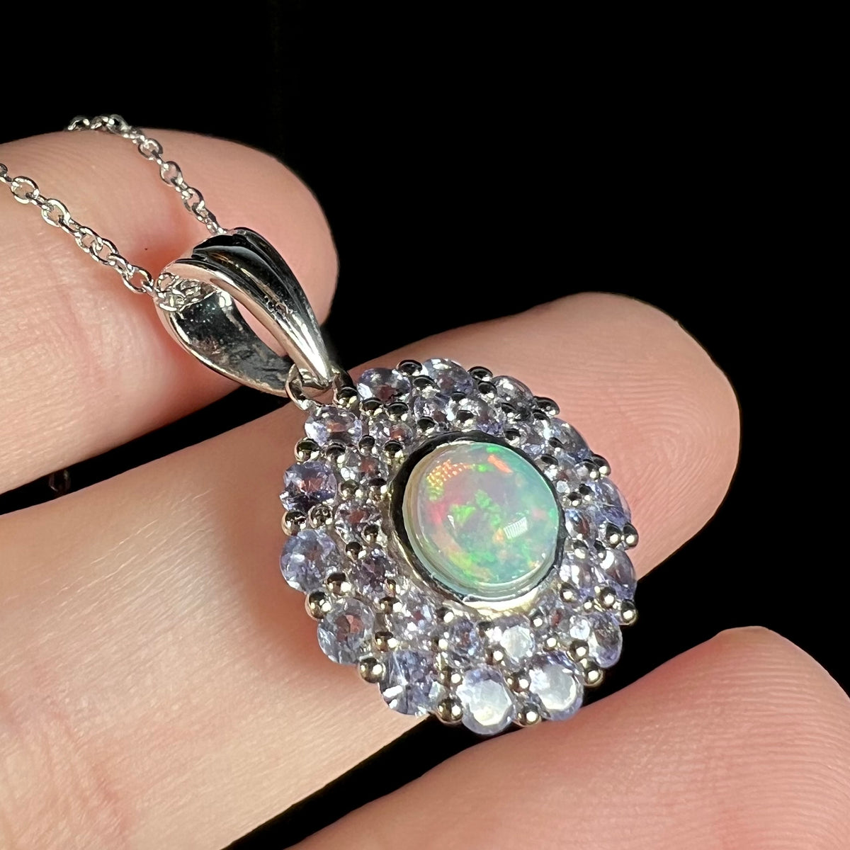 Angled close-up of a sterling silver opal and tanzanite double halo pendant, shown resting on fingers beneath cool LED studio light.