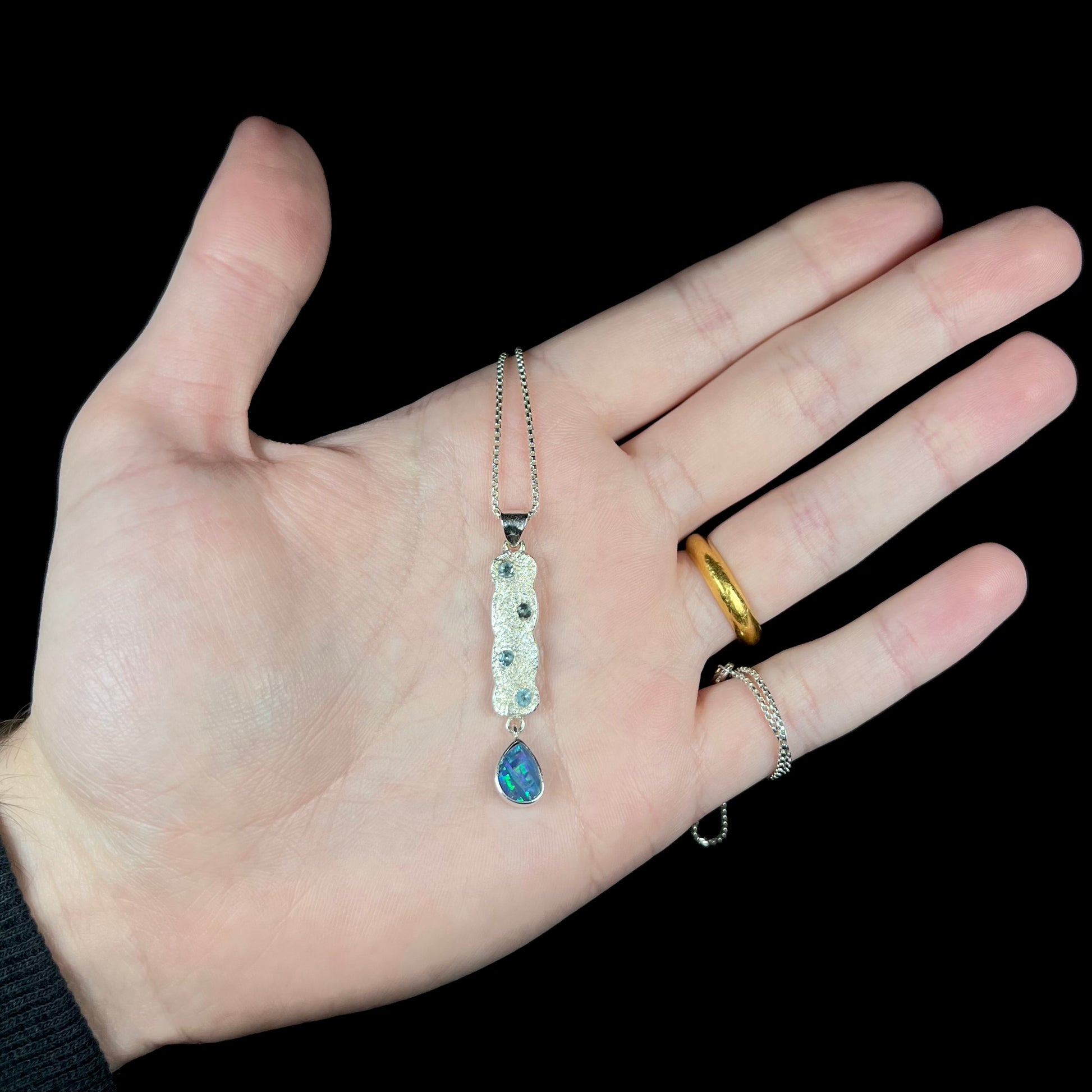 An estate sterling silver Australian opal doublet and blue topaz drop necklace held in a hand under cool LED light against black background.