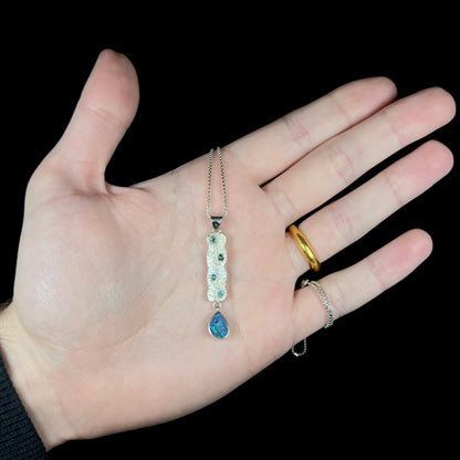 An estate sterling silver Australian opal doublet and blue topaz drop necklace held in a hand under cool LED light against black background.