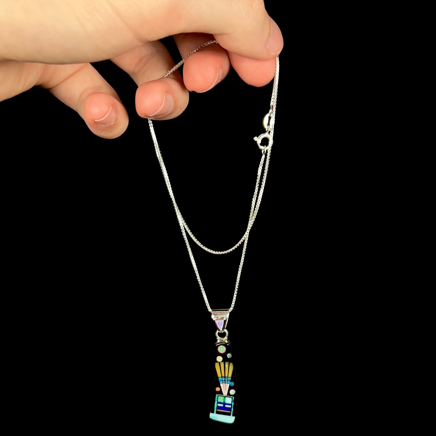 Hand dangling sterling silver Southwest-style inlay necklace from its chain against black background.