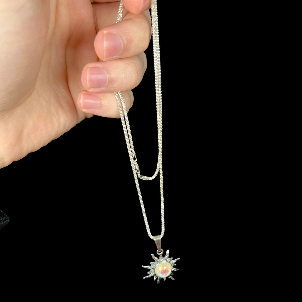 Hand dangling a sterling silver imitation opal sun necklace from its silver chain.