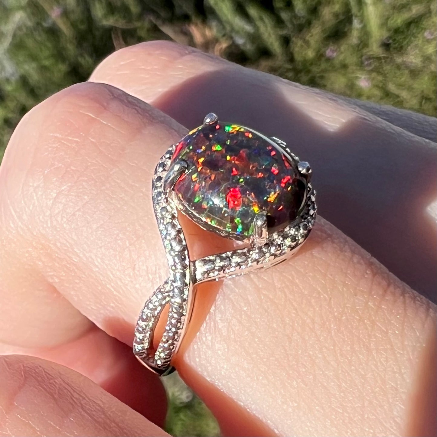 A lab-created black opal solitaire ring with red color play in sterling silver, shown on a finger in sunlight at an angle.