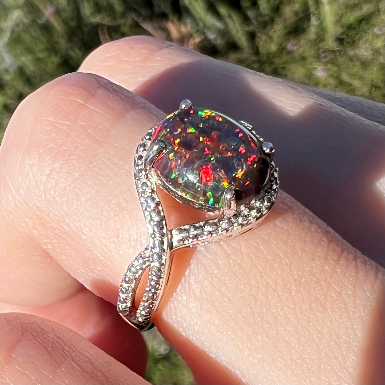 A lab-created black opal solitaire ring with red color play in sterling silver, shown on a finger in sunlight at an angle.