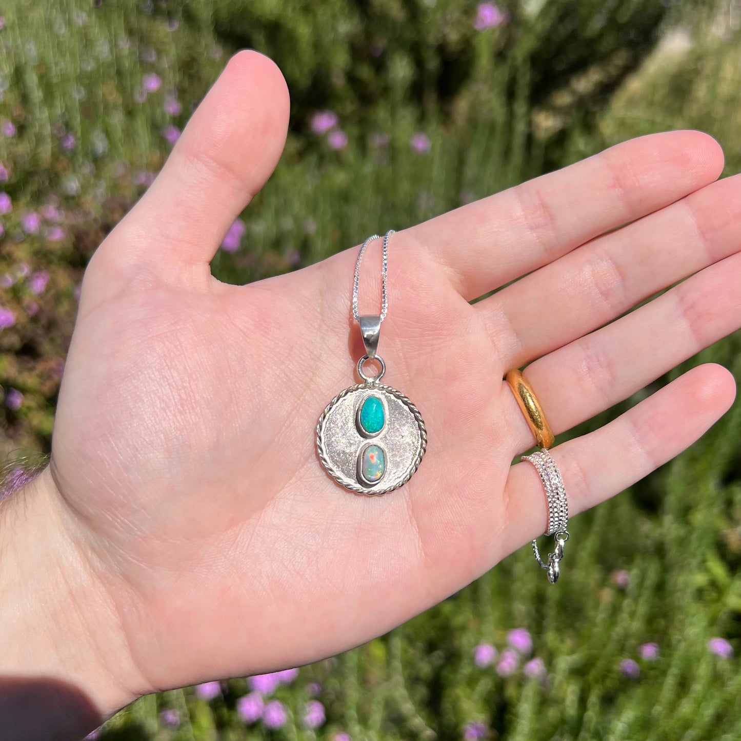 A round, quarter-sized sterling silver two-stone Australian opal men's pendant held in a hand in the sunlight.  One opal is blue-green, the other has red flashes.