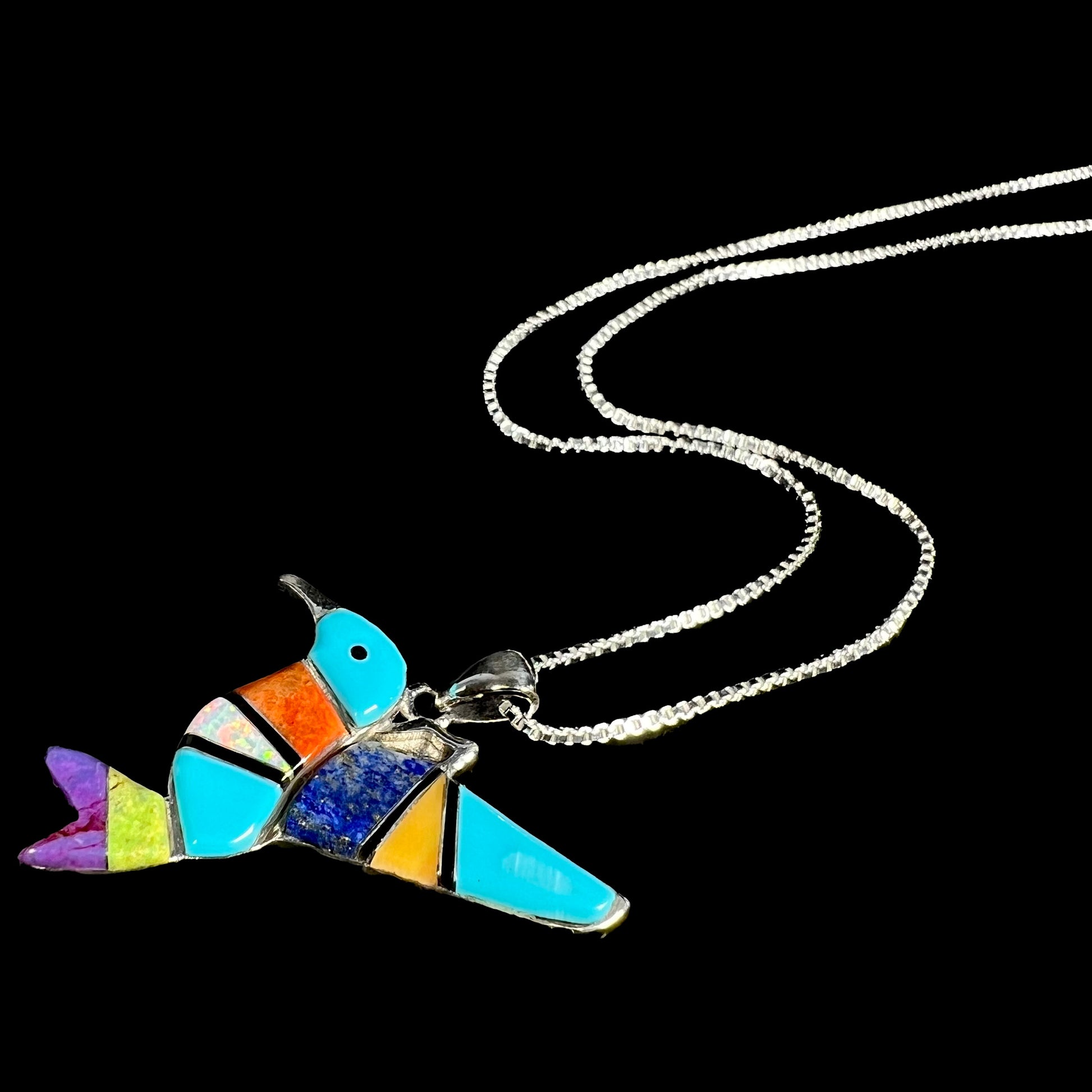 A multi-stone inlay hummingbird necklace in sterling silver, shown at an angle, laying against a black backdrop.