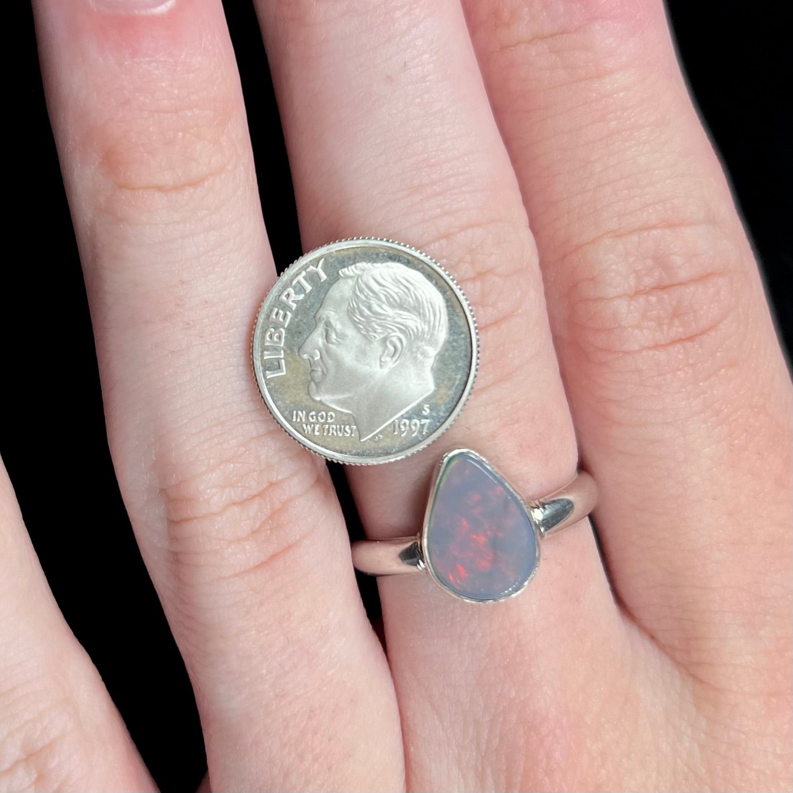 A silver pear shaped Australian opal solitaire ring is worn on a hand and shown next to a US dime coin for scale.  The opal has subtle red flashes.