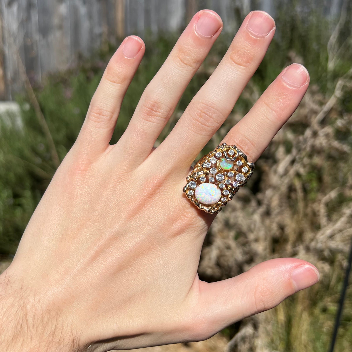 Hand wearing a two-stone synthetic opal ring with many CZ accents in natural sunlight.