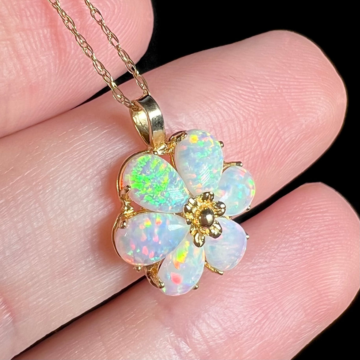 Close-up angled view of a synthetic opal flower pendant in 14k gold, held in a hand and shown on a gold chain