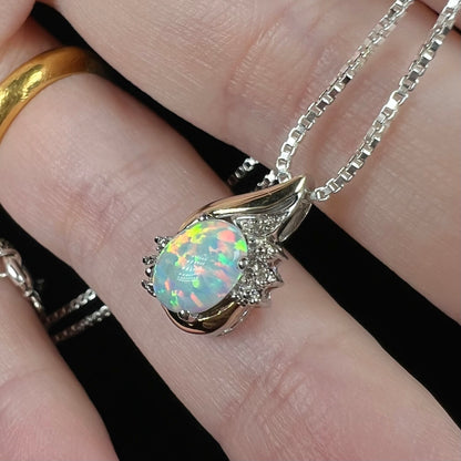 A close-up of a silver chain necklace with an oval-shaped synthetic opal pendant, held on a person's fingers. The pendant features a vibrant, iridescent opal center stone surrounded by silver settings and small diamond accent stones.
