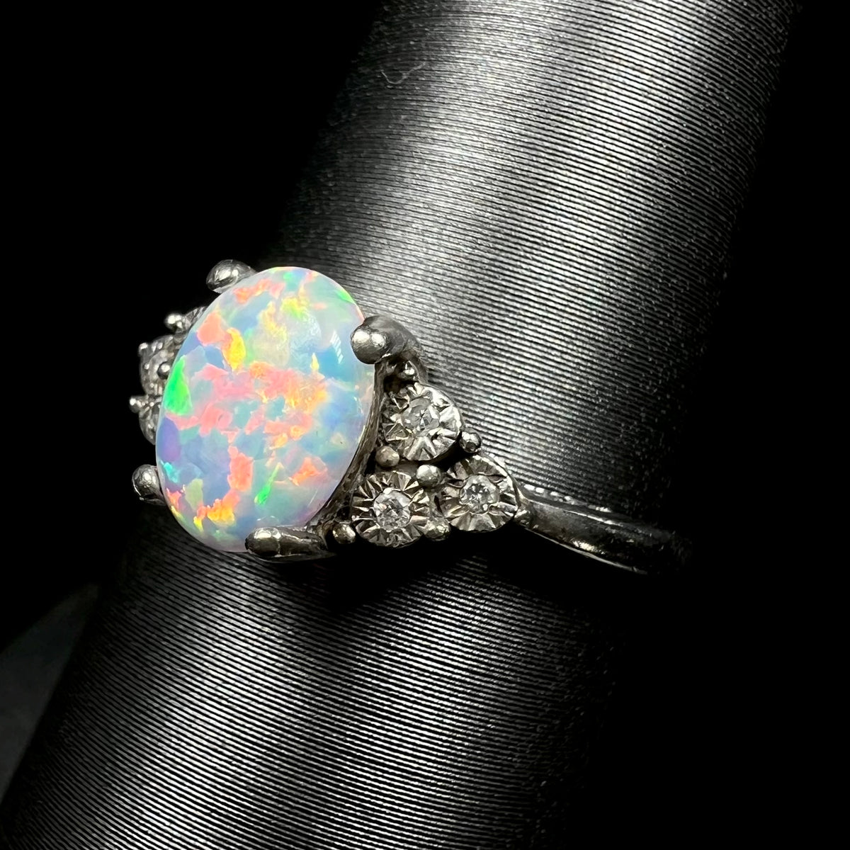 Angled shoulder view of synthetic opal ring focusing on illusion set natural diamond accents, shown on ring display.  The opal has a bright red flash of color.