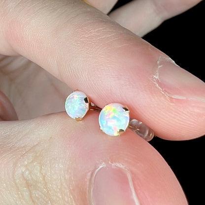 Angled close-up view of a pair of synthetic opal stud earrings set in solid 14k yellow gold prongs, held between two fingers under cool LED studio light.
