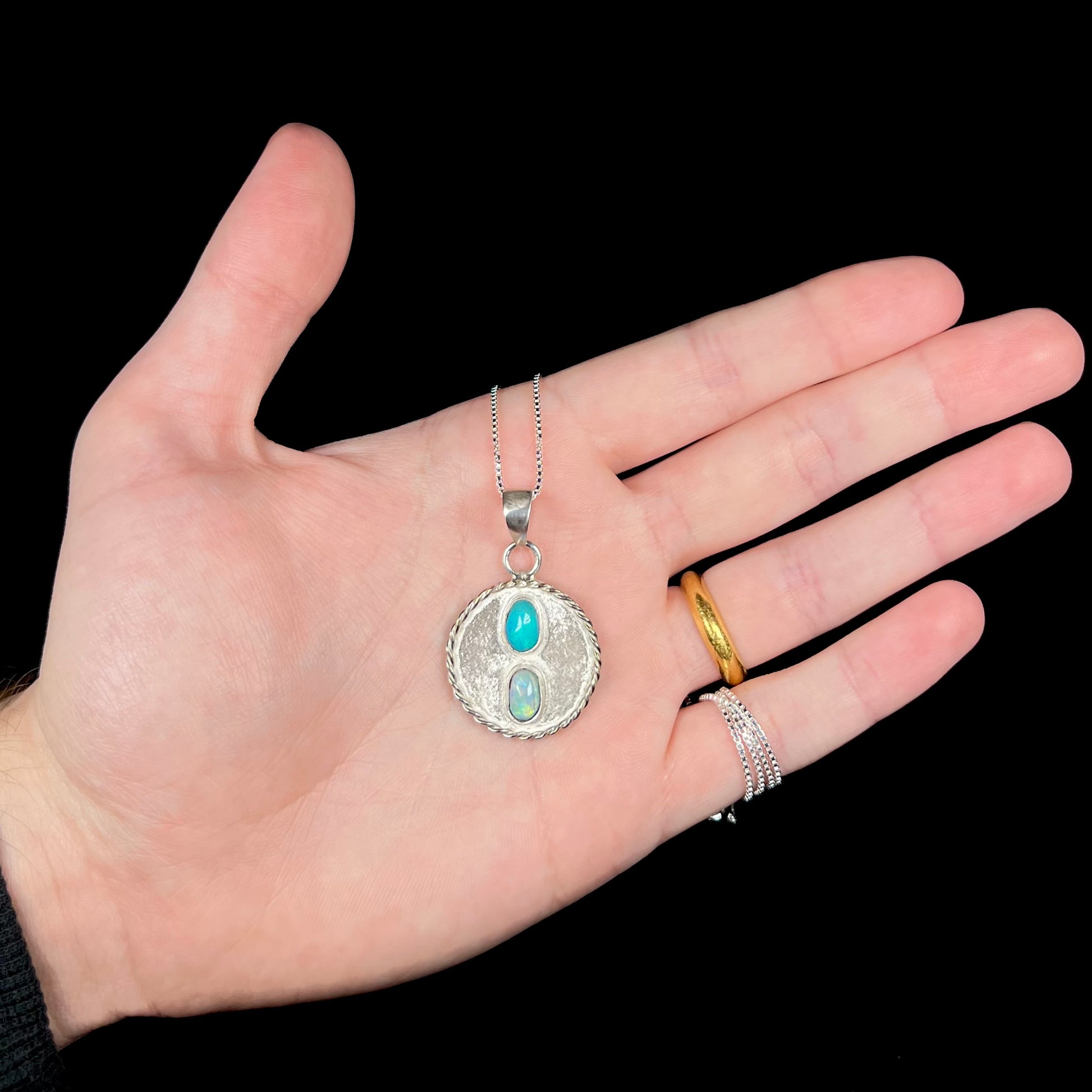 Hand holding a quarter-sized textured sterling silver men's opal necklace against black background under LED light.