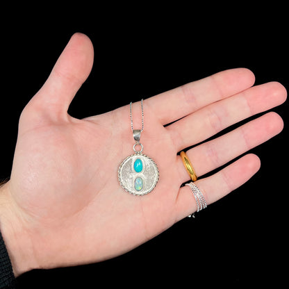 Hand holding a quarter-sized textured sterling silver men's opal necklace against black background under LED light.
