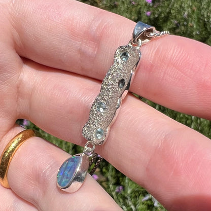 Close-up side view of a sterling silver opal drop pendant with a textured design, shown in natural sunlight.