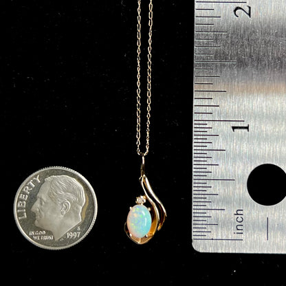 A delicate gold opal and diamond pendant measuring three-quarters of an inch in length, shown between a US dime coin and ruler