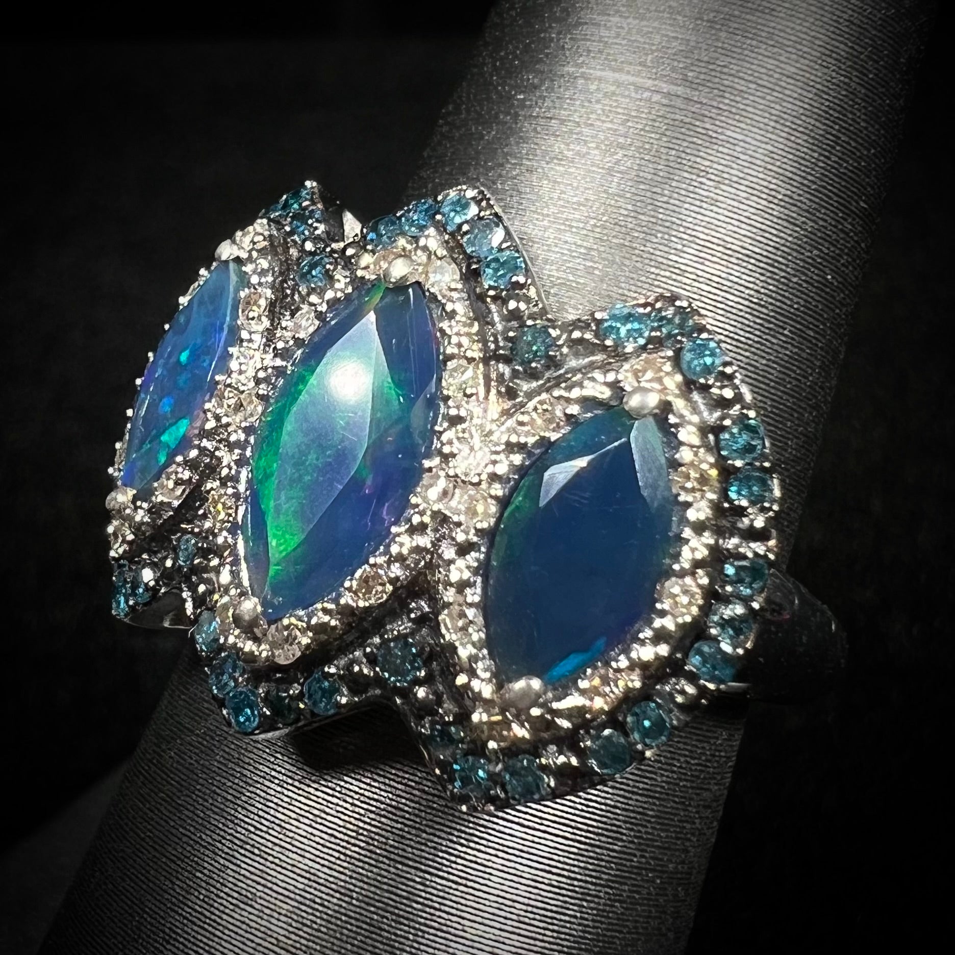 A three-stone blue opal and diamond halo ring, shown at an angled view against a black backdrop.