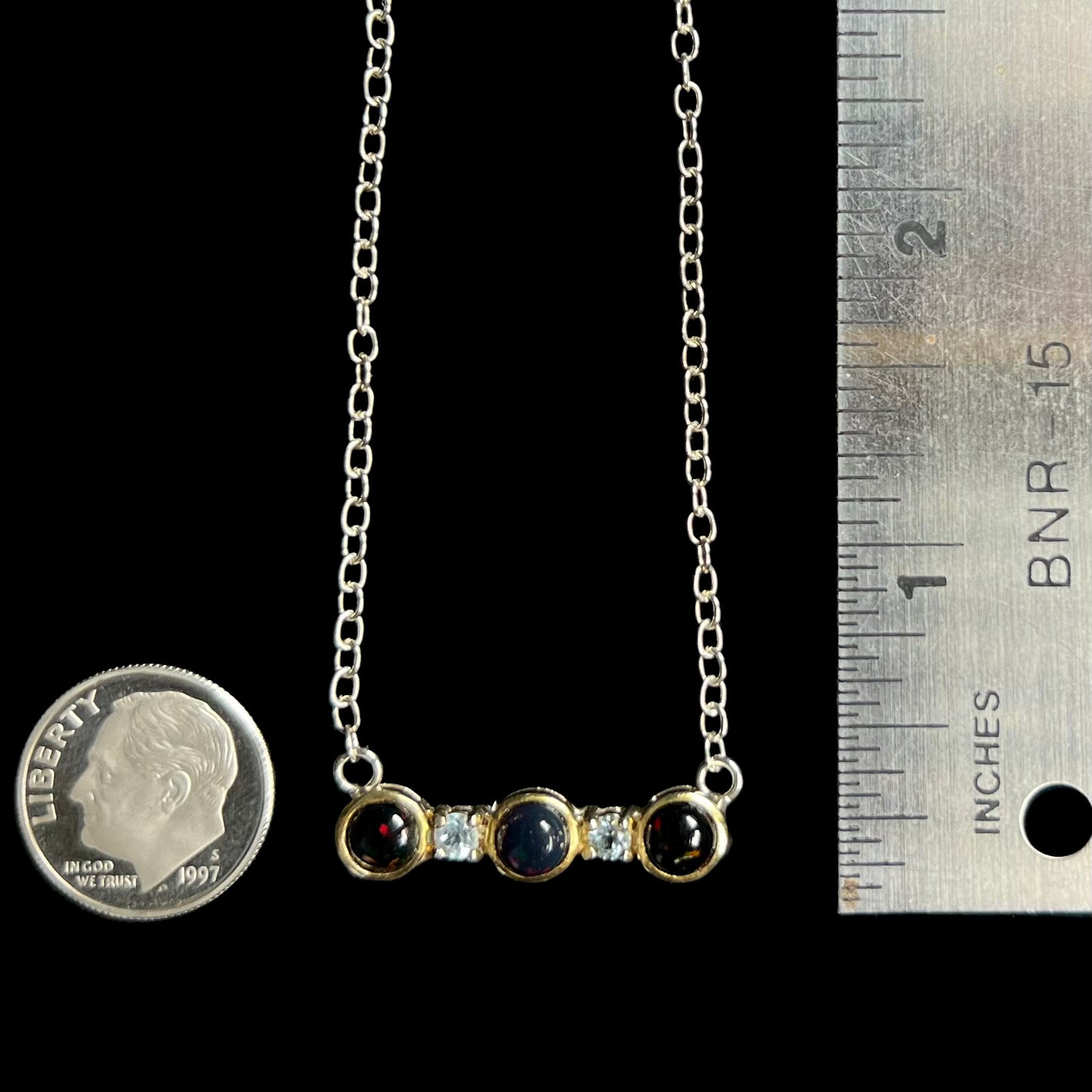 A three-stone black Ethiopian opal and white topaz necklace in two-tone sterling silver, next to a dime and ruler.
