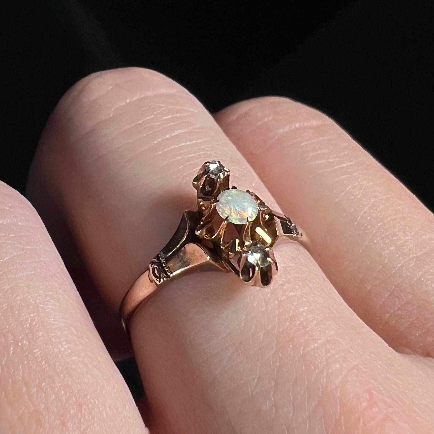 Three-stone opal and rose cut diamond ring shown close-up from an angle on a finger against a dark backdrop.