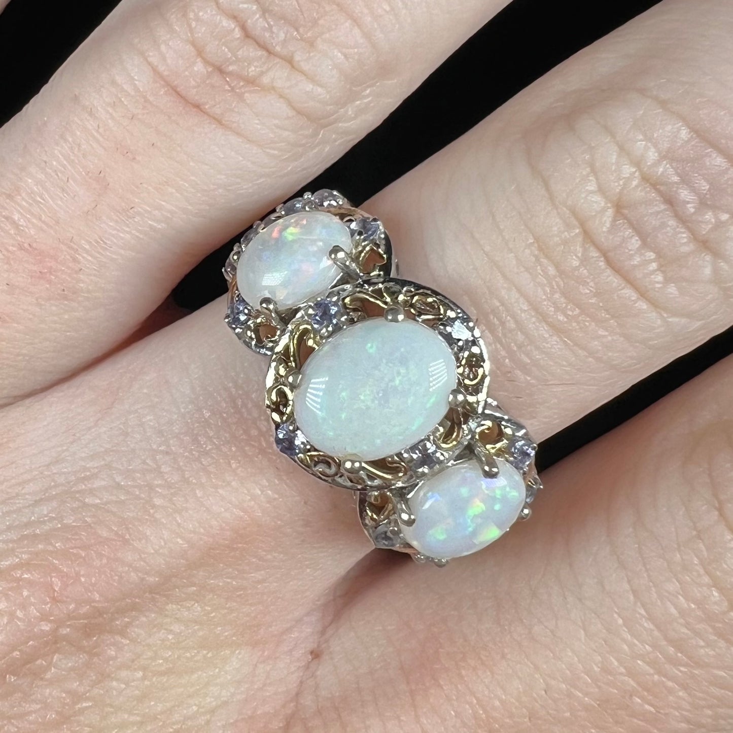 Close-up of a three-stone Australian opal ring with a halo of open gold-tone filigree and tanzanite accents, shown on a finger.