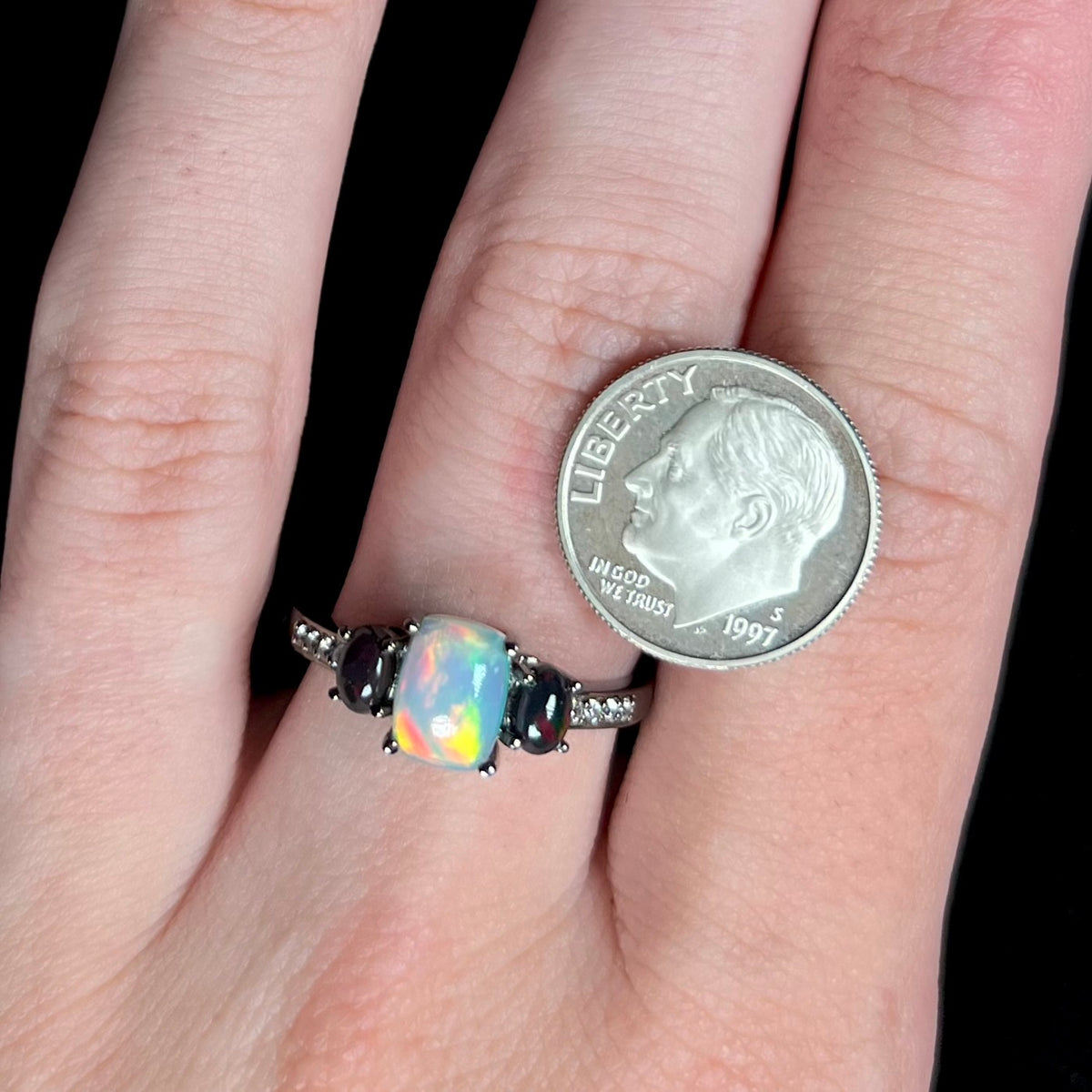 Close-up of hand wearing three-stone opal and CZ ring with black rhodium finish, shown next to US dime coin