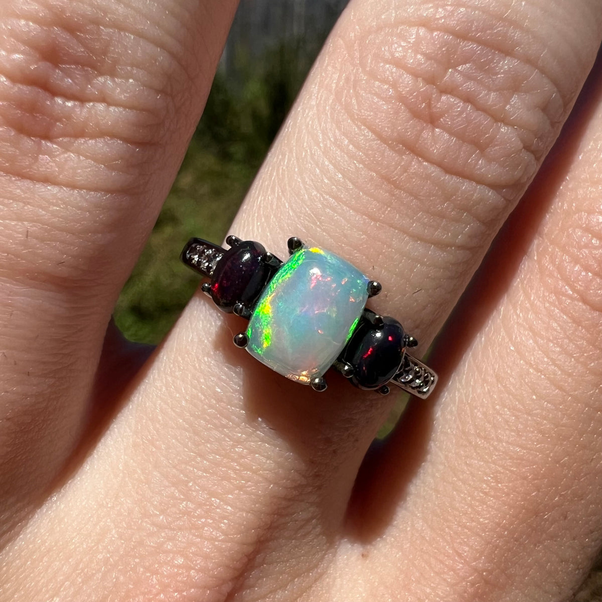 Close-up sunlit view of three-stone Ethiopian opal ring with black rhodium finish, worn on finger