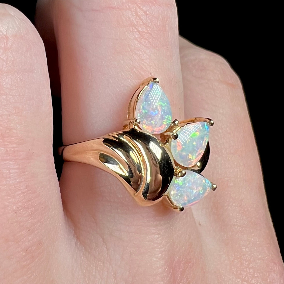 Yellow gold three-stone opal ring shown close-up from angle, worn on finger.  The opals are pear shaped, tips pointing outward.