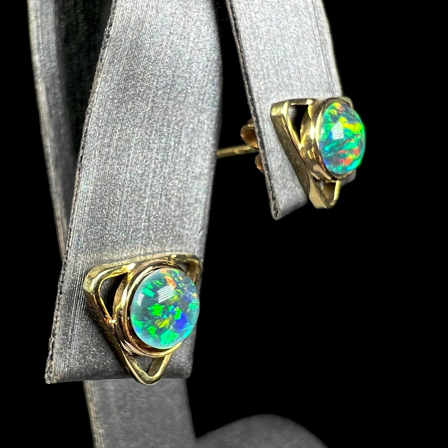 Angled close-up of gold triangle stud earrings set with round black opal triplets.