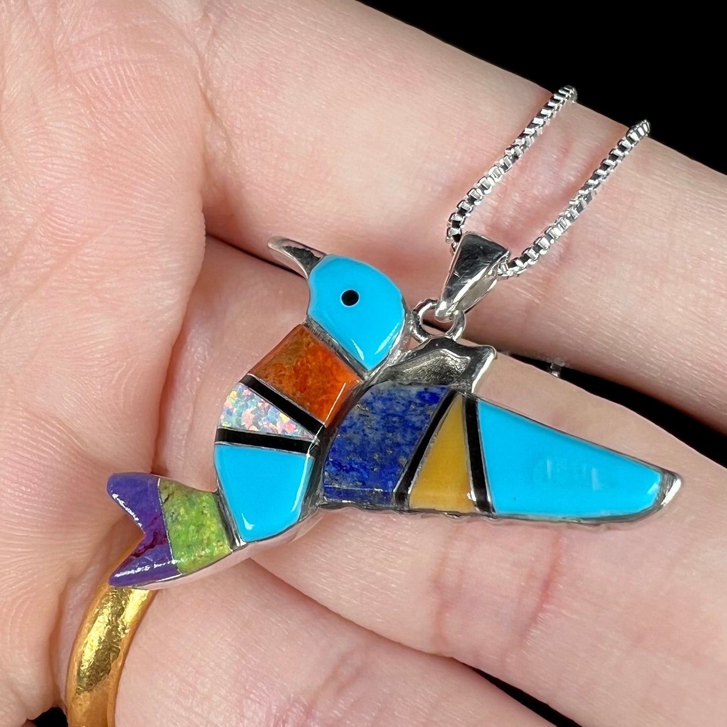 A multi-stone inlay hummingbird necklace in sterling silver, shown at an angle close-up in a hand.