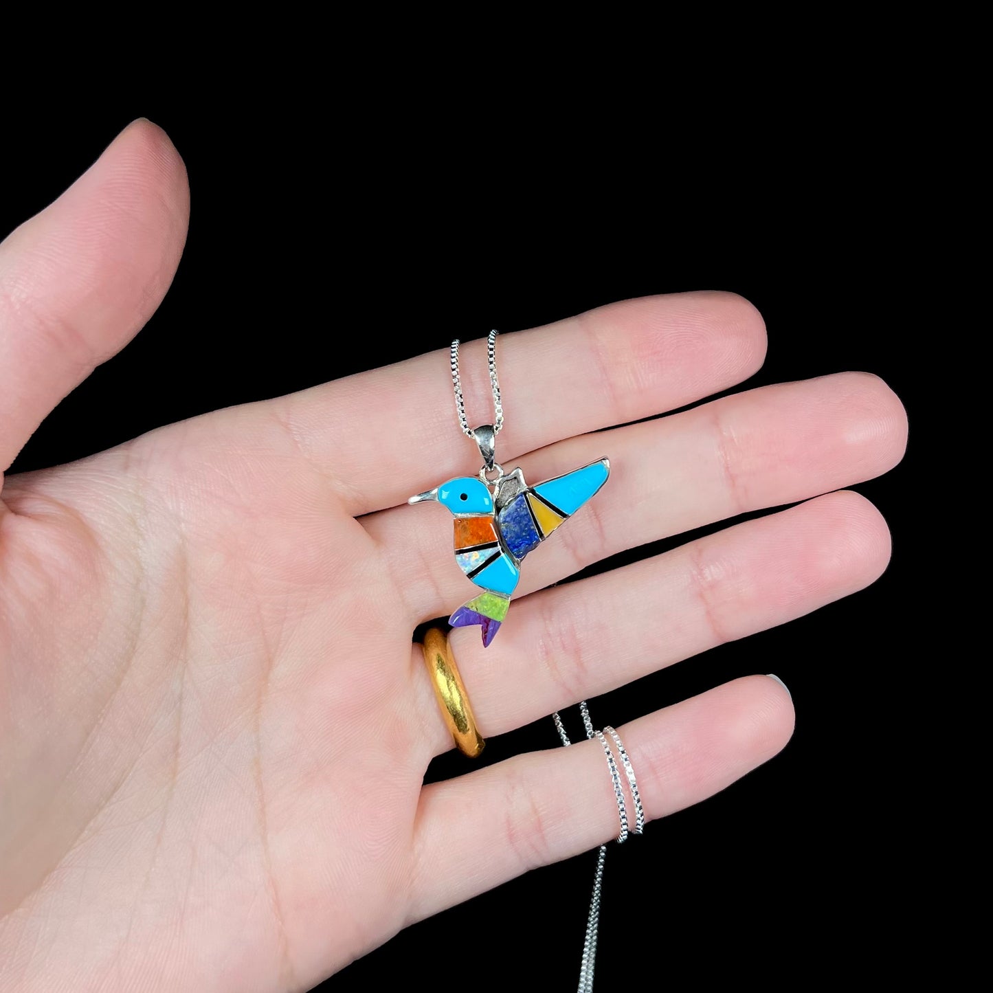 A multi-stone inlay hummingbird necklace in sterling silver, shown in a hand against a black background.