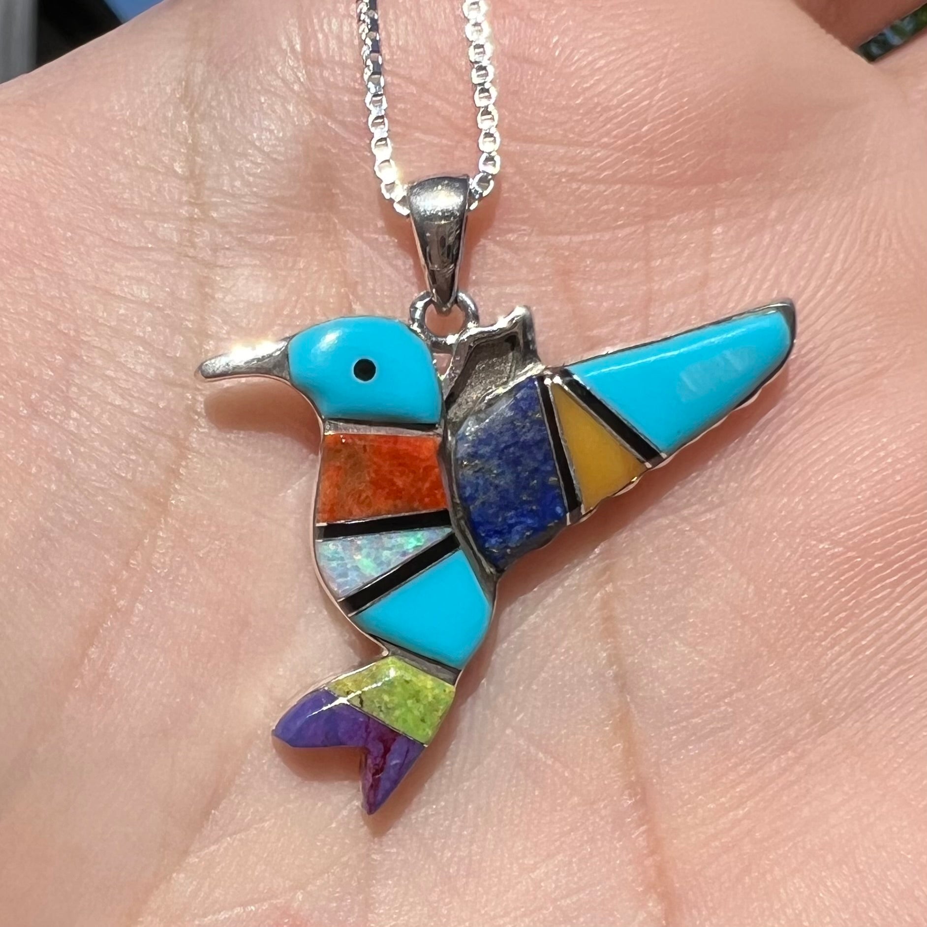A multi-stone inlay hummingbird necklace in sterling silver, shown close-up in a hand in sunlight.
