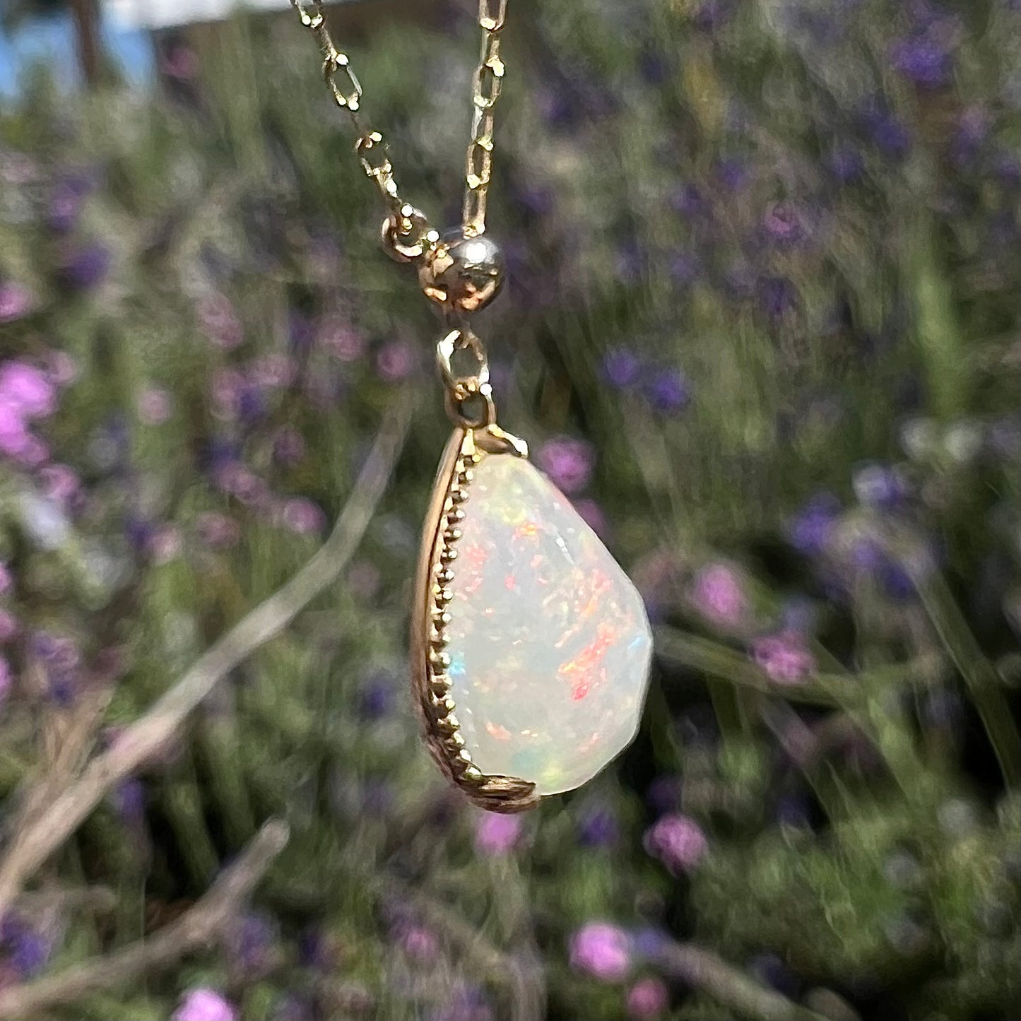 Macro view of the opal necklace at a twisted angle in sunlight, showing faint red flashes and small prongs securing the opal. Green foliage and purple flowers in the background.