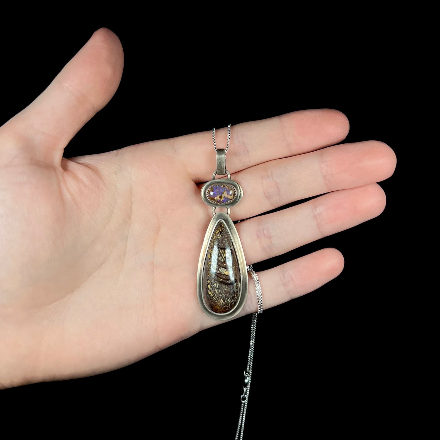 Hand holding a silver two-stone boulder opal pendant on a box chain against a black background under cool studio light.