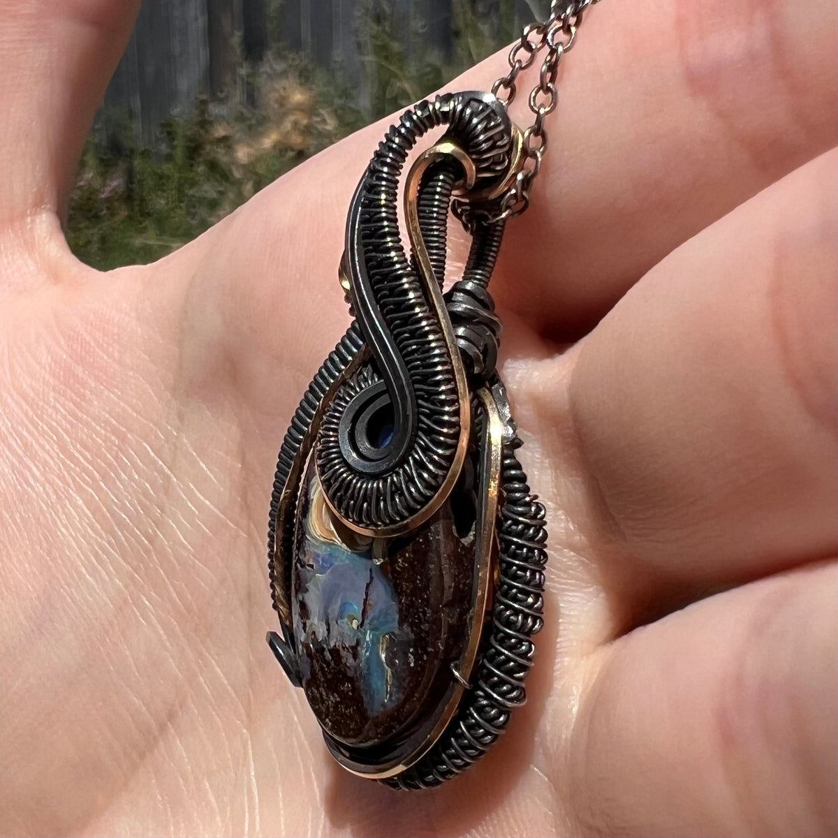 A two-tone antiqued sterling silver and gold-plated wire wrapped Australian boulder opal pendant shown close-up from an angled view in a hand, held in sunlight.