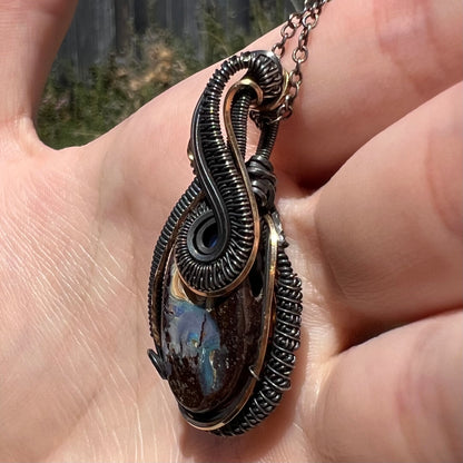 A two-tone antiqued sterling silver and gold-plated wire wrapped Australian boulder opal pendant shown close-up from an angled view in a hand, held in sunlight.