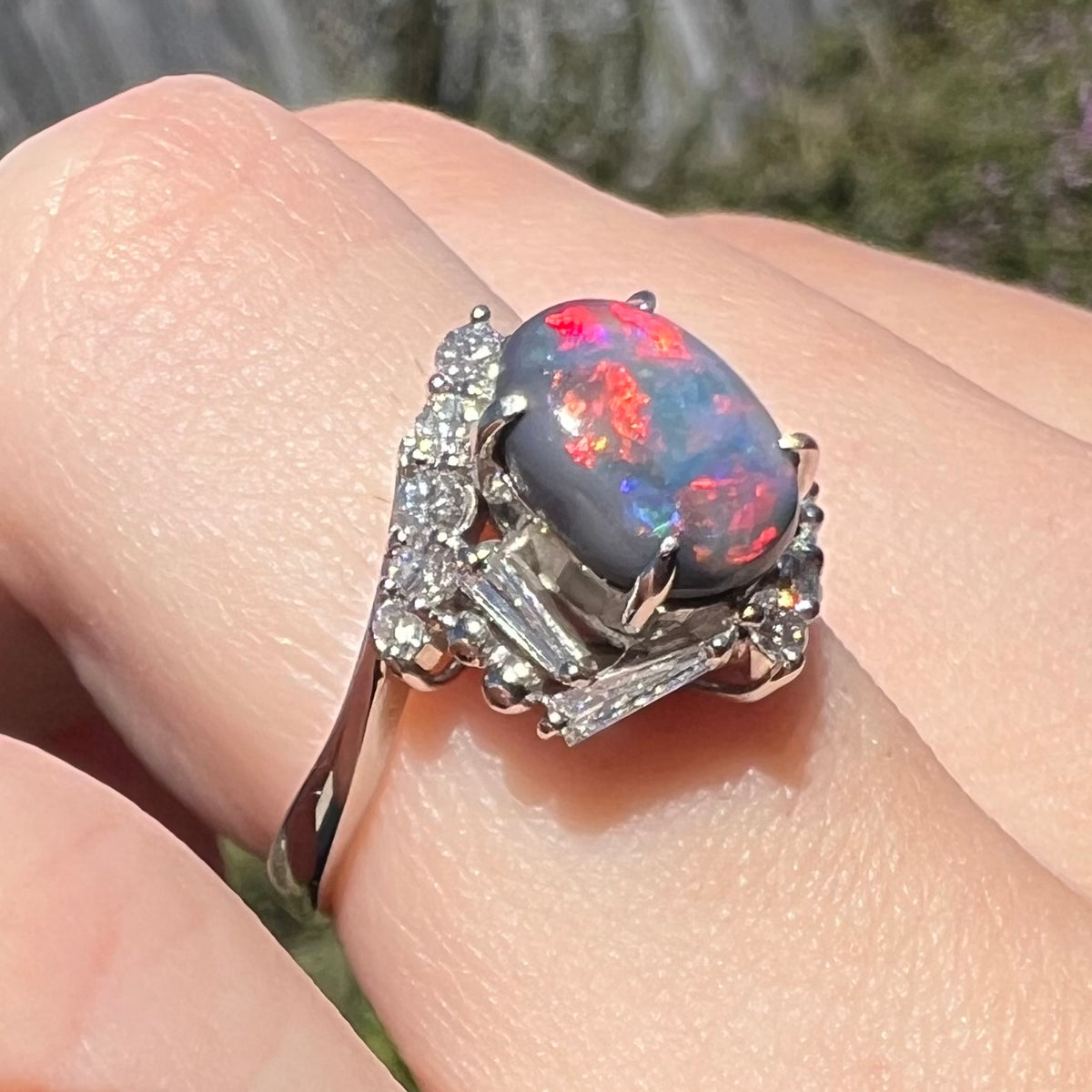 A unique platinum opal and diamond engagement ring viewed on finger close-up from angle, red play-of-color seen over the opal's N5-dark, periwinkle-colored body.