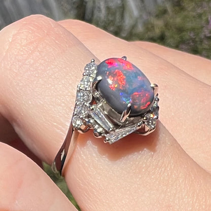 A unique platinum opal and diamond engagement ring viewed on finger close-up from angle, red play-of-color seen over the opal's N5-dark, periwinkle-colored body.
