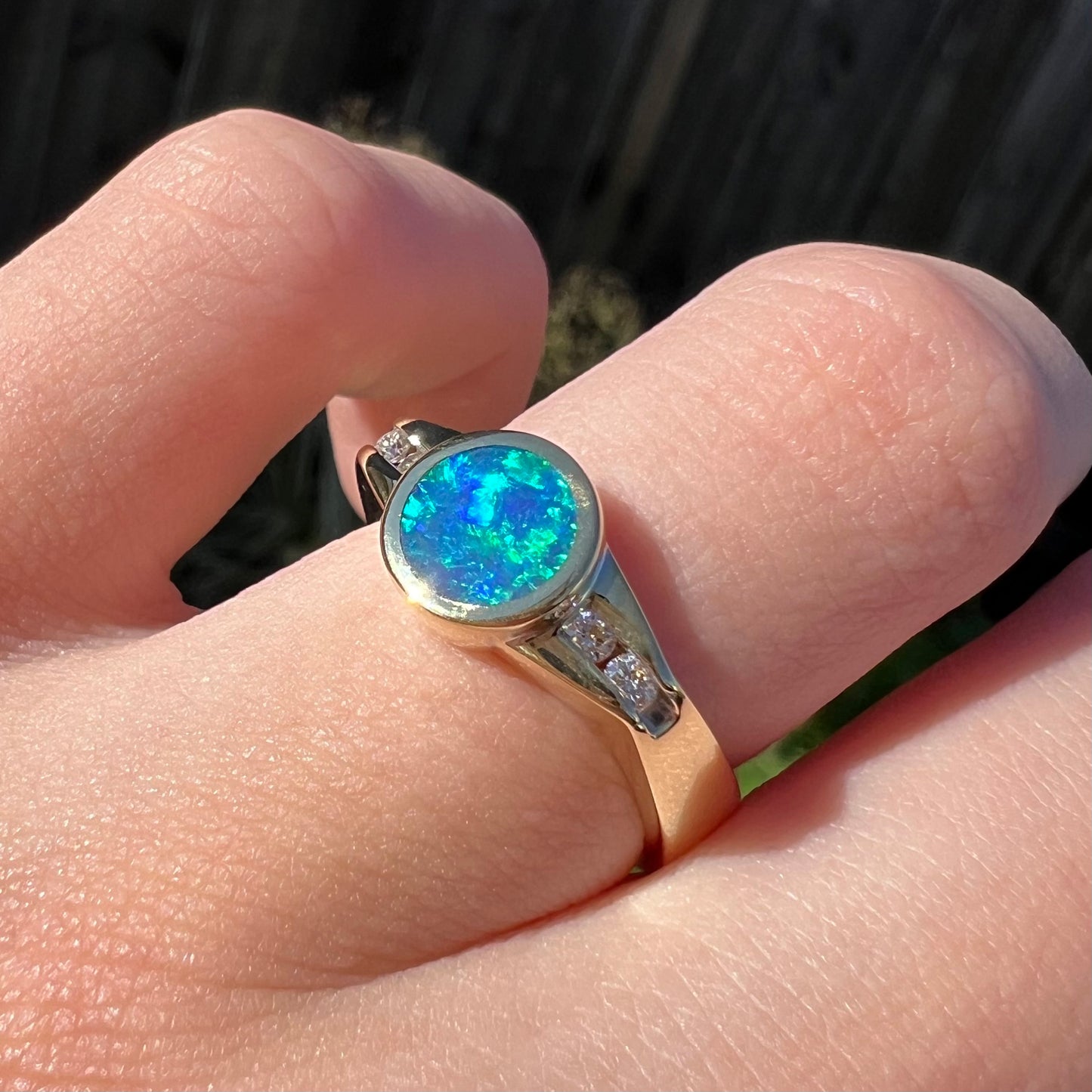 Angled close-up of a unisex black boulder opal and diamond ring worn on a male's finger in the sunlight against a wooden fence background.