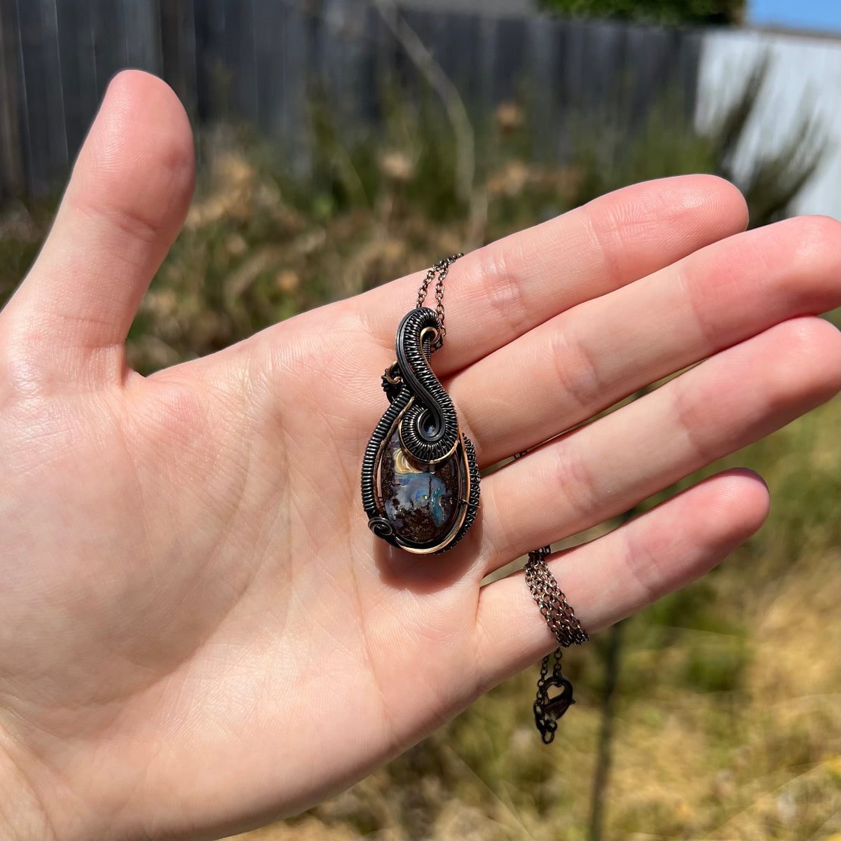 Hand holding a unisex two-tone blackened sterling silver and gold-tone wire wrapped boulder opal pendant in sunlight.
