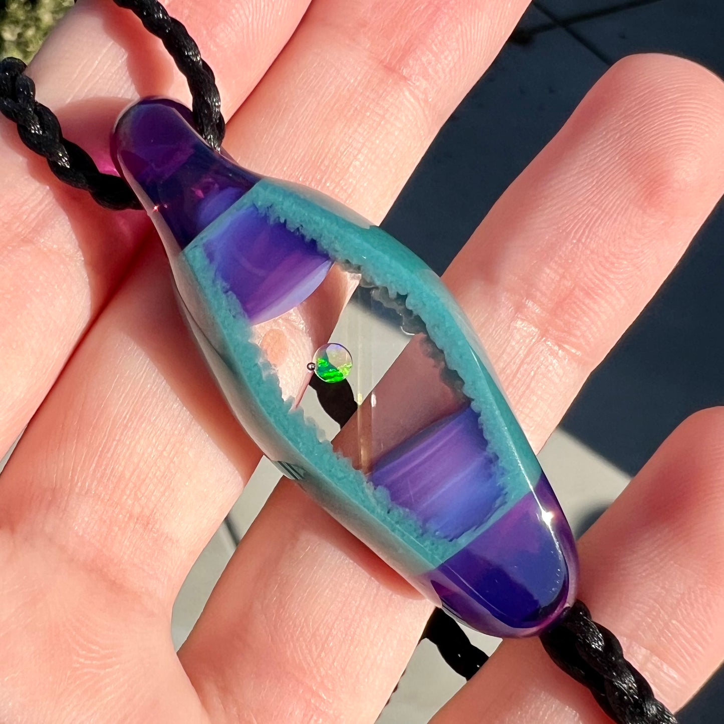 Blue and purple unisex boro glass and opal pendant held in the sunlight by a hand, shown close-up from an angle.