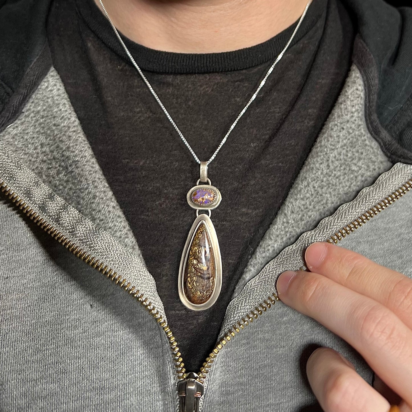 Man wearing a unisex sterling silver boulder matrix opal necklace around his neck inside of a hoodie against a dark shirt.