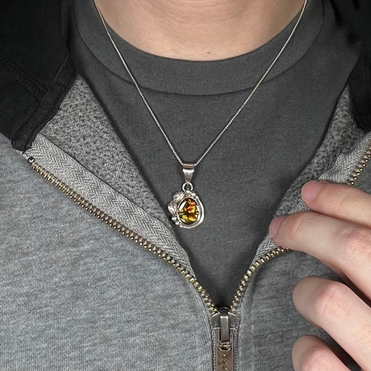 Close-up of person's neck and chest region wearing unisex fire agate necklace in sterling silver against dark shirt beneath partially unzipped hoodie.
