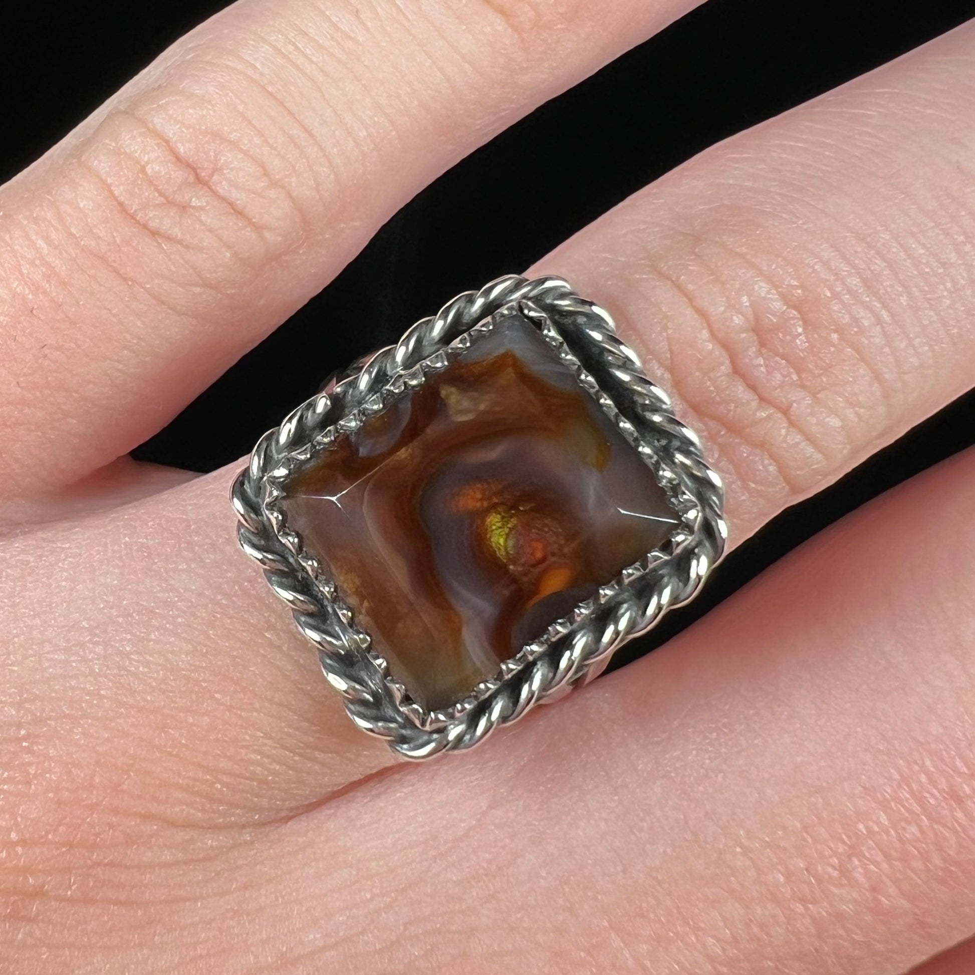 Finger wearing a unisex fire agate ring with silver rope design against black backdrop.