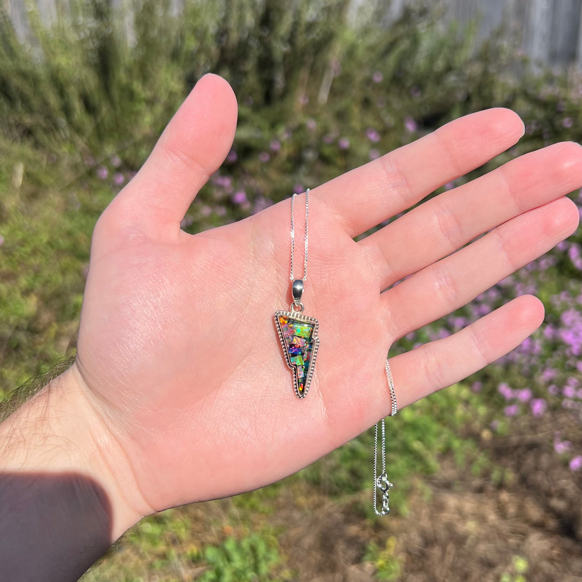 Sunlit hand holding unisex mosaic opal lightning bolt necklace against blurred outdoor background.