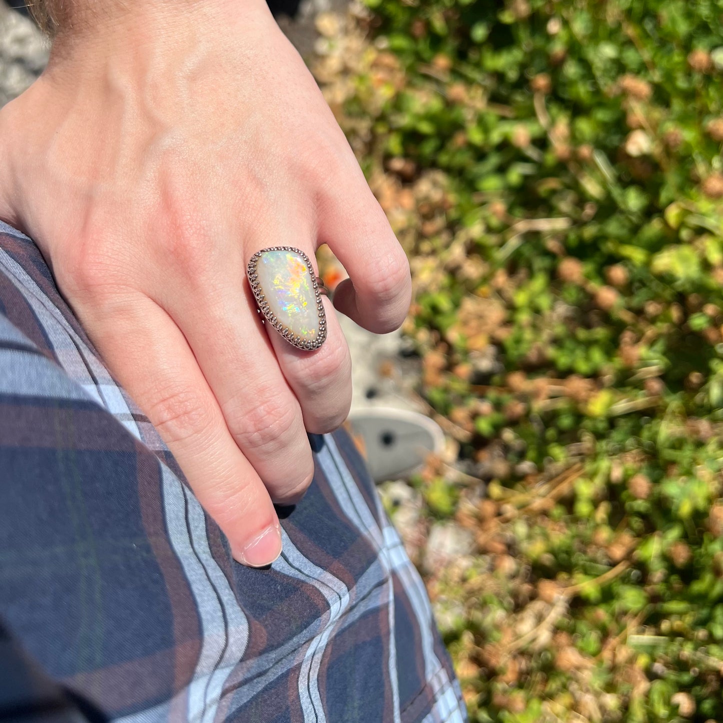 A unisex natural opal solitaire ring worn on a hand as the hand rests on a person's hip in sunlight against a blurred green background.