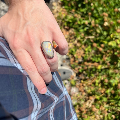 A unisex natural opal solitaire ring worn on a hand as the hand rests on a person's hip in sunlight against a blurred green background.