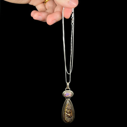 Unisex sterling silver Koroit bouulder opal necklace dangled by a hand from its box style silver chain against black background.